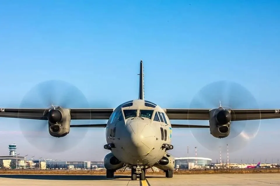 Un avion militar cu patru motoare cu elice este poziționat pe o pistă de aterizare sub un cer senin.