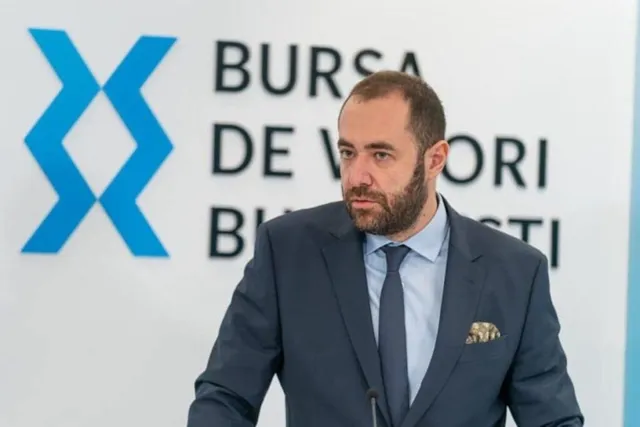 Un bărbat cu barbă poartă un costum închis la culoare, cămașă albastră și cravată, stând în fața unui panou alb cu text și un simbol albastru.
