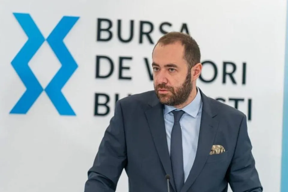 Un bărbat cu barbă poartă un costum închis la culoare, cămașă albastră și cravată, stând în fața unui panou alb cu text și un simbol albastru.