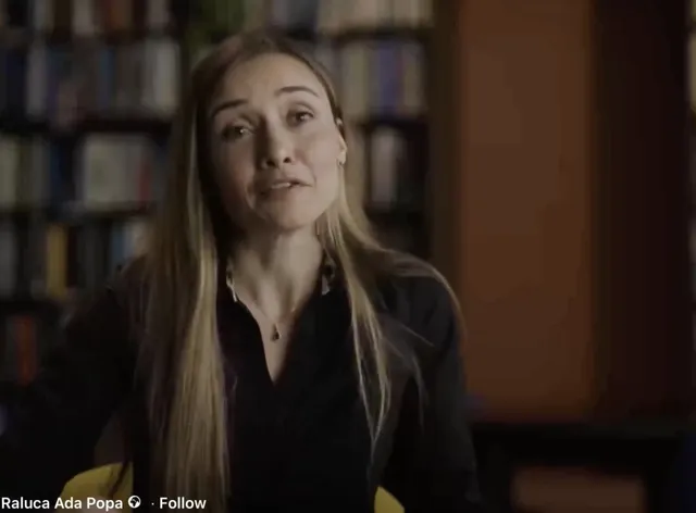 O femeie cu părul lung și deschis la culoare poartă o cămașă neagră și un colier subțire, stând în fața unui fundal cu rafturi de cărți.