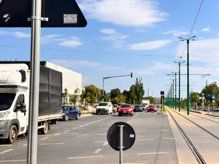 Mașini circulă pe un drum asfaltat lângă șine de tramvai, sub un cer senin cu nori albi și lângă un camion alb.