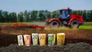 Mai multe bancnote de euro sunt înfipte vertical în pământ, în prim-plan, în fața unui câmp agricol cu un tractor roșu în fundal.