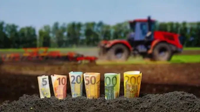 Mai multe bancnote de euro sunt înfipte vertical în pământ, în prim-plan, în fața unui câmp agricol cu un tractor roșu în fundal.