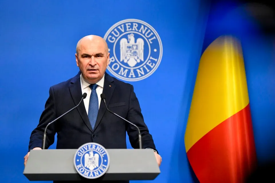 Un bărbat cu părul scurt și costum închis stă în spatele unui podium cu sigiliul Guvernului României, lângă un steag tricolor.
