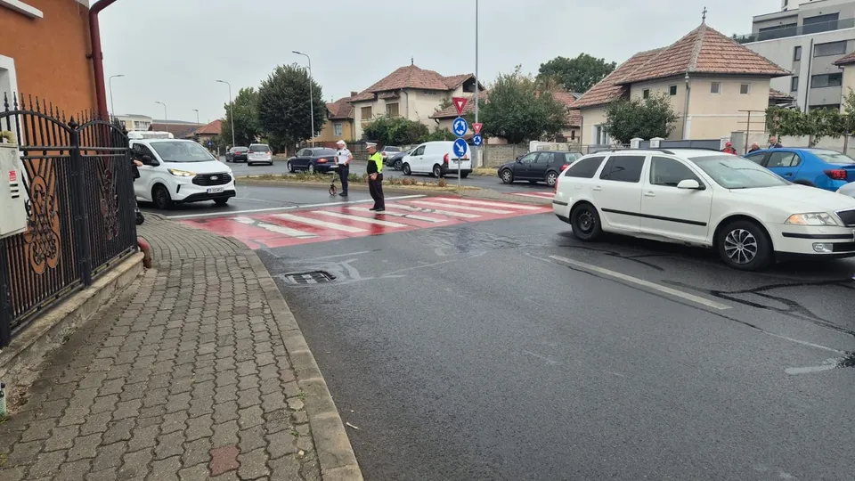 Două persoane stau pe o trecere de pietoni marcate cu linii albe, în timp ce mai multe autoturisme circulă pe un drum asfaltat lângă o zonă rezidențială.
