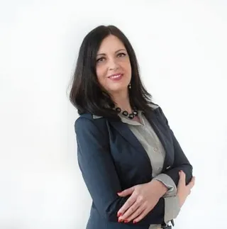 O femeie cu părul negru, purtând un sacou albastru și o bluză gri, stă cu brațele încrucișate în fața unui fundal alb.