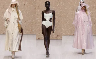 Trei modele merg pe o pistă de catwalk purtând haine de culoare crem, alb și roz pal, incluzând o rochie de baie și două trenchuri.
