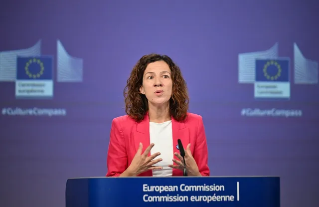 O femeie cu părul creț, purtând un sacou roz și un tricou alb, vorbește în spatele unui podium albastru cu textul Comisiei Europene.