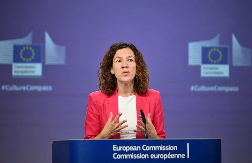 O femeie cu părul creț, purtând un sacou roz și un tricou alb, vorbește în spatele unui podium albastru cu textul Comisiei Europene.