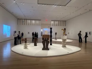 Oamenii vizitează diverse sculpturi de culoare bronz și alb pe platforme albe într-o sală de muzeu cu pereți albi și podea de lemn.