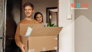 Un bărbat zâmbind poartă o cutie de carton deschisă, în timp ce o femeie stă în spatele lui într-o intrare de apartament.