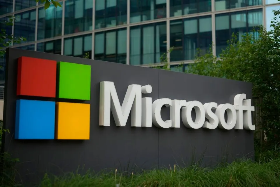 Un semn cu logo colorat și textul alb Microsoft este montat pe un panou gri în fața unei clădiri de birouri cu ferestre de sticlă.