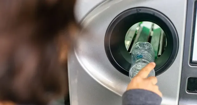 O persoană ține o sticlă de plastic transparentă în fața unei deschideri circulare de pe un aparat metalic de colectare.