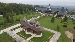 O biserică de lemn cu trei turnuri este situată pe un teren verde, înconjurată de alei de piatră și copaci.