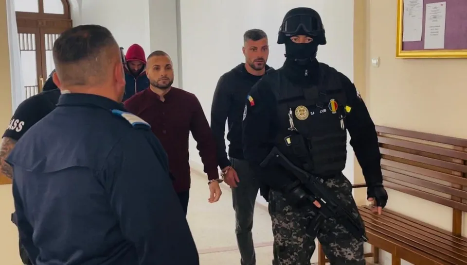 Un agent de poliție în echipament tacticos negru și cască stă în prim-plan, în fața a doi bărbați care merg într-un coridor lângă un banc de lemn.
