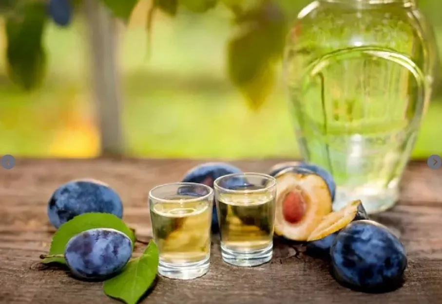 Două pahare mici cu lichid transparent sunt așezate pe o suprafață de lemn lângă câteva fructe de prune și o sticlă de sticlă.