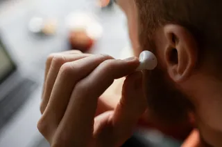 O persoană ține un dispozitiv audio mic și alb lângă urechea dreaptă într-un cadru cu fundal neclar.