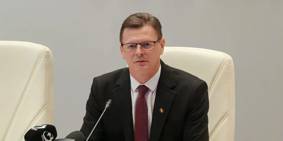 Un bărbat cu ochelari, purtând sacou negru, cămașă albă și cravată purpurie, stă așezat într-un scaun alb lângă un microfon.