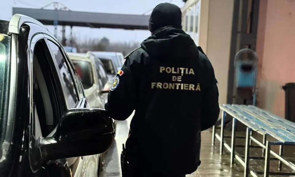 Un agent de poliție cu uniformă neagră stă întors, lângă un vehicul negru și o bancă metalică, într-un punct de control.