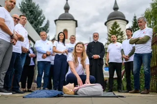 Un grup de oameni stă în picioare pe o esplanadă lângă un manechin de antrenament întins pe o saltea sub cerul înnorat.