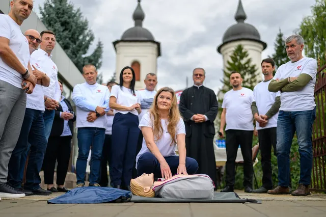 Un grup de oameni stă în picioare pe o esplanadă lângă un manechin de antrenament întins pe o saltea sub cerul înnorat.