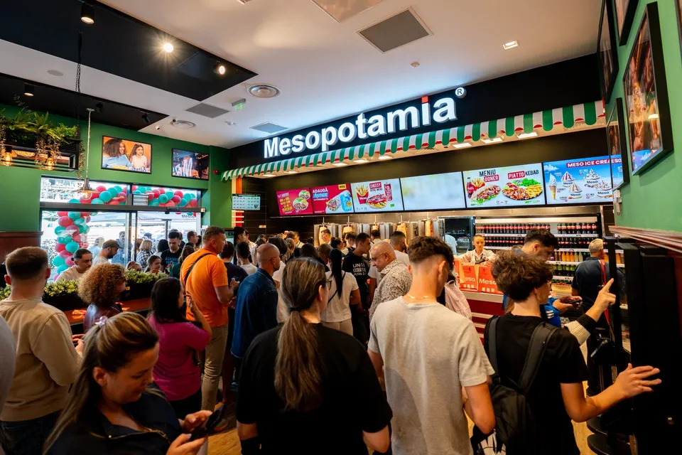O mulțime de oameni stă în coadă în interiorul unui restaurant, lângă un tejghea cu meniuri afișate și băuturi răcoritoare.