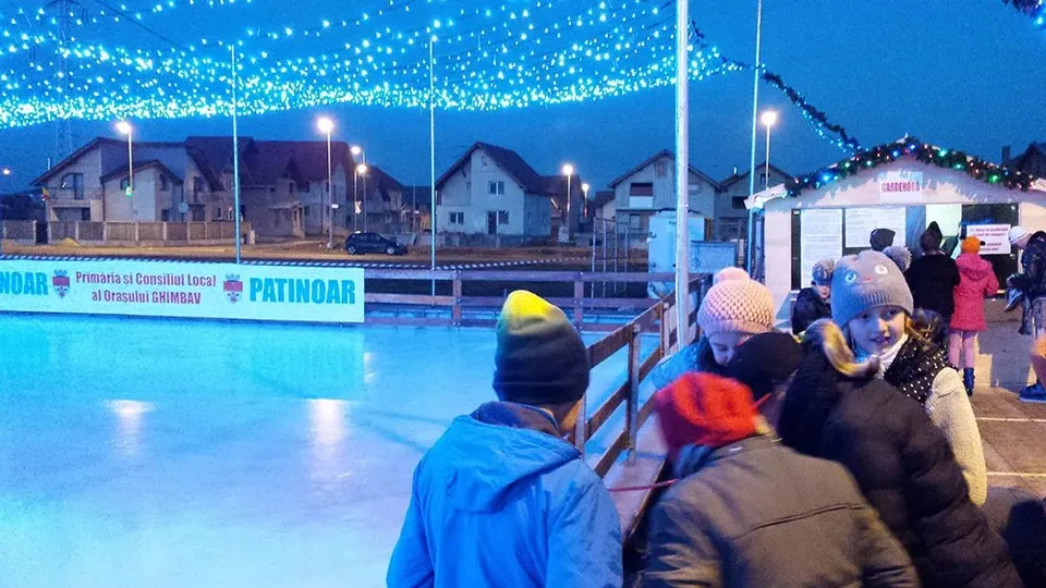 Oamenii stau pe marginea unei suprafețe de gheață lângă un banner albastru și alb, sub un decor de lumini verzi suspendate.