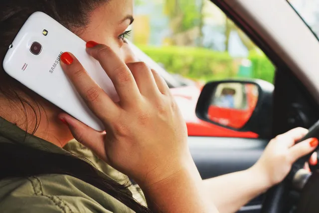 O femeie cu unghiile vopsite în roșu ține un telefon mobil alb la ureche în timp ce conduce o mașină.