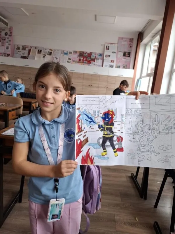 O fată cu o cămașă albastră și un ID la gât ține un desen colorat cu un pompier, lângă o carte de colorat în deschidere.