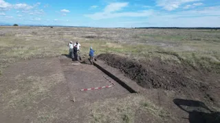 Patru persoane stau lângă o groapă de excavare într-un câmp deschis sub un cer cu nori dispersate.