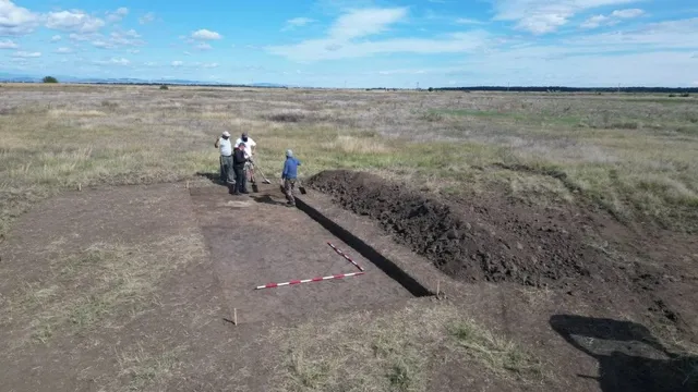 Patru persoane stau lângă o groapă de excavare într-un câmp deschis sub un cer cu nori dispersate.