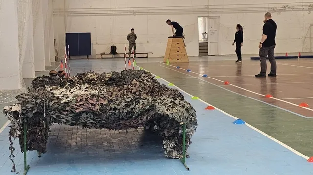 O plasă de camuflaj întinsă pe podeaua unei săli de sport, în timp ce câțiva oameni se află în fundal lângă echipamente de antrenament.