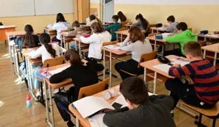 Elevi stau la bănci în interiorul unei săli de clasă, scriind în caiete și lucrând cu materiale de scris pe mesele de lemn.