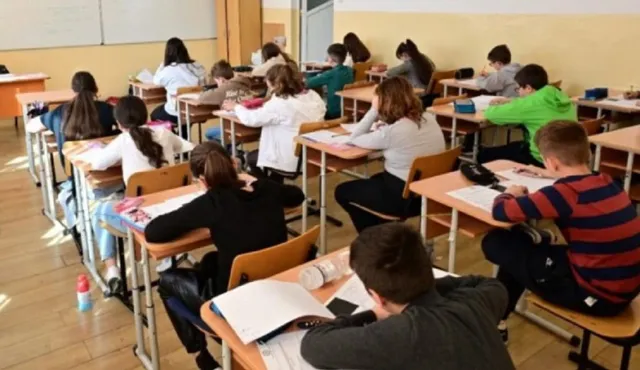 Elevi stând la birouri de clasă, unii scriind în caiete, alții folosind tabletele sau telefoanele, într-o sală de cursă cu tablă și ferestre.