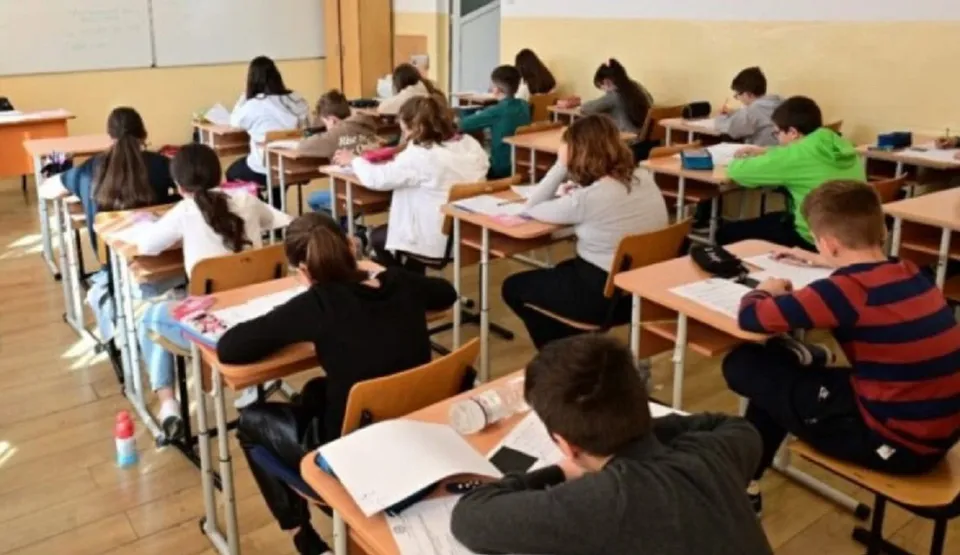 Elevi stând la birouri de clasă, unii scriind în caiete, alții folosind tabletele sau telefoanele, într-o sală de cursă cu tablă și ferestre.