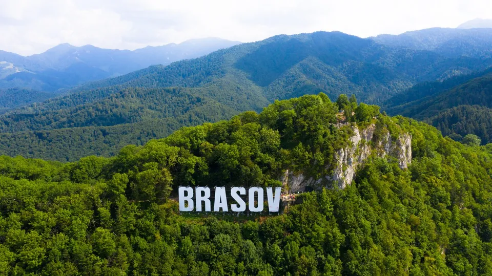 Un semn alb cu textul BRASOV este amplasat pe o colină împădurită, sub un perete stâncos, având munți în fundal.