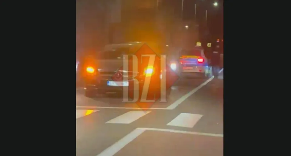 Două autoturisme sunt parcate pe un drum asfaltat cu marcaje de trecere de pietoni, sub o lumină portocalie intensă, pe timp de noapte.