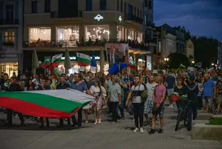 O mulțime de oameni merge pe o stradă pietonală purtând diverse steaguri, inclusiv unul mare cu dungi verzi, albe și roșii.