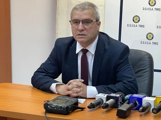 Un bărbat cu ochelari, purtând sacou închis la culoare, cămașă albă și cravată purpurie, stă la o masă cu mai multe microfoane în față.