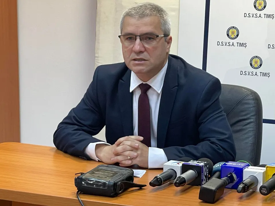 Un bărbat cu ochelari, purtând sacou închis la culoare, cămașă albă și cravată purpurie, stă la o masă cu mai multe microfoane în față.