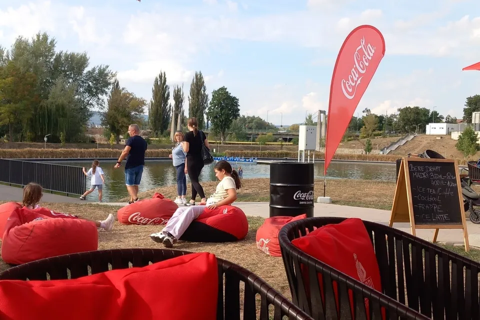 Oamenii stau pe pufuri roșii lângă un lac, în timp ce un steag Coca-Cola și un panou de meniu sunt vizibile în fundal.