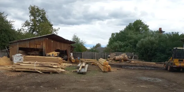 Stive de scânduri de lemn sunt depozitate pe un teren deschis lângă un șopron de lemn sub un cer înnorat.