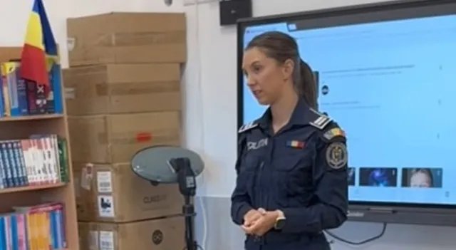 O femeie în uniformă albastră stă în fața unui ecran mare, având cutii de carton și rafturi cu cărți în fundal.