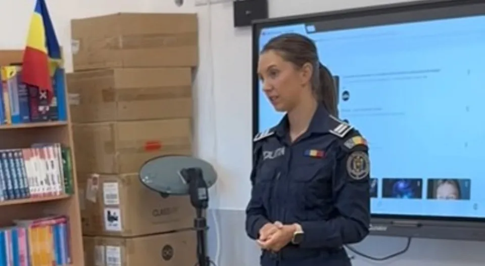 O femeie în uniformă albastră stă în fața unui ecran mare, având cutii de carton și rafturi cu cărți în fundal.
