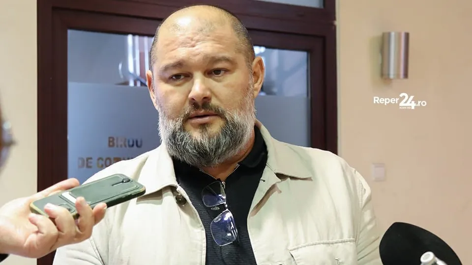 Un bărbat cu barbă și ochelari agățați de cămașă poartă o jachetă deschisă, fiind încadrat de microfoane și un telefon mobil.
