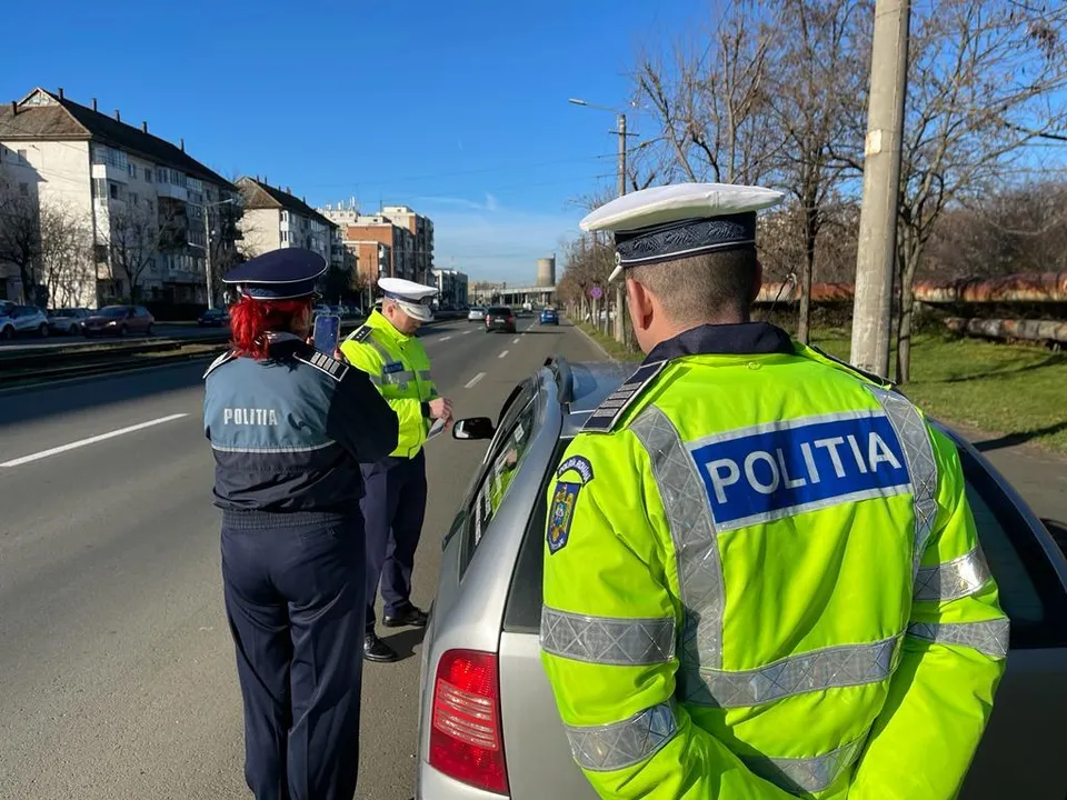 Trei polițiști în uniforme de serviciu, inclusiv unul cu vestă reflectorizantă galbenă, stau lângă o mașină gri pe marginea unei drumuri asfaltate.