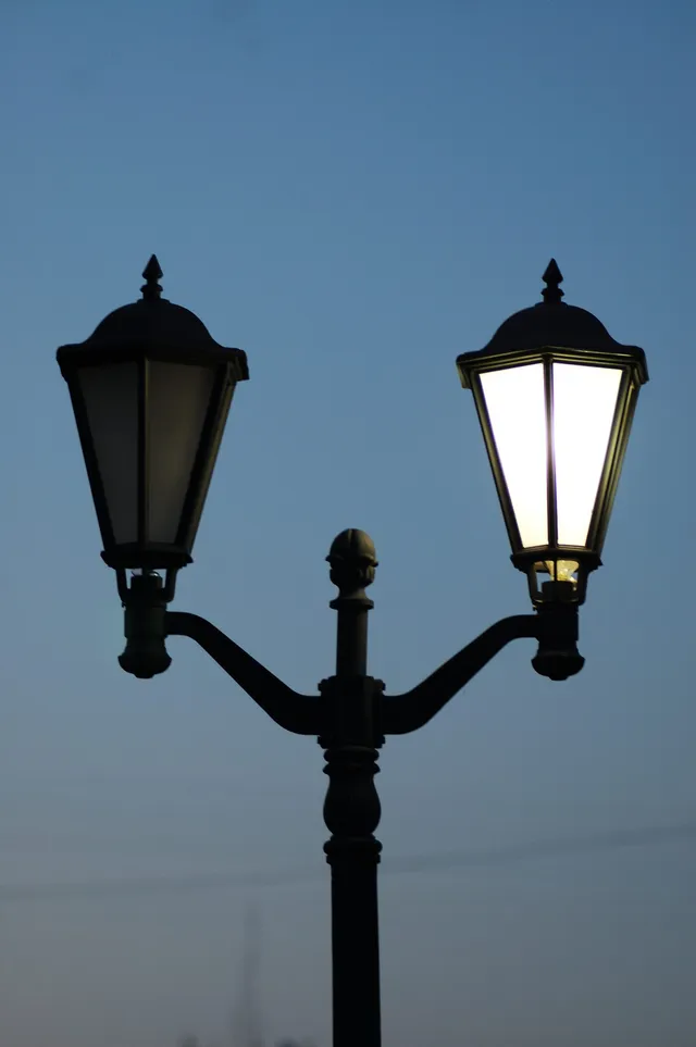 Un stâlp de iluminat public cu două corpuri de lumină, unul aprins și unul stins, sub un cer de seară.
