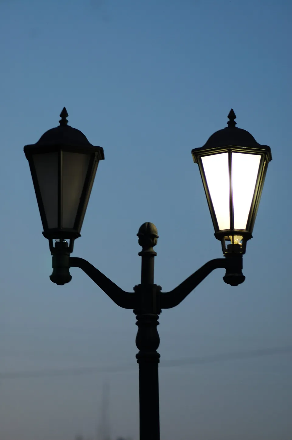 Un stâlp de iluminat public cu două corpuri de lumină, unul aprins și unul stins, sub un cer de seară.