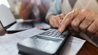 Mâini apasă tastele unui calculator de birou așezat peste documente, lângă un laptop, într-un cadru interior neclar.