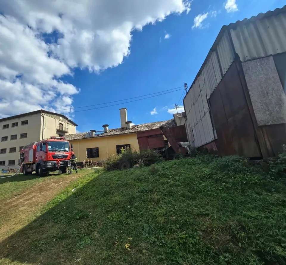 Un camion de pompieri roșu este parcat pe un teren verde lângă o clădire cu acoperiș de tablă sub un cer cu nori albi.
